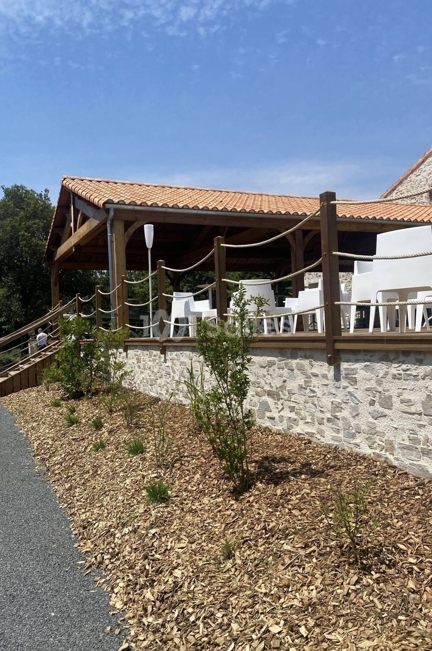 Terrasse couverte en bois avec toiture en tuiles, chaises blanches et cordes décoratives, vue sur bâtiment en pierre.