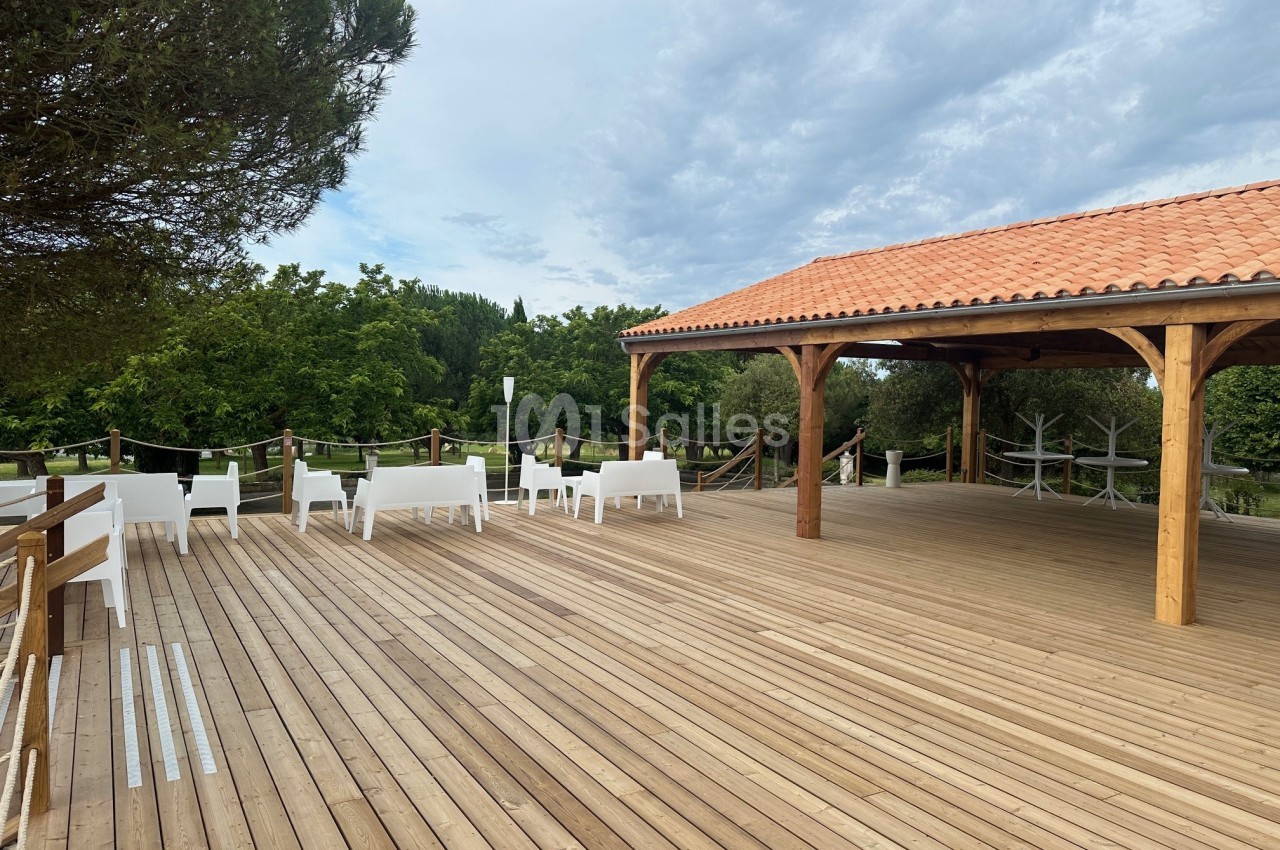 Terrasse en bois avec chaises blanches, structure couverte en bois et tuiles, entourée de verdure.