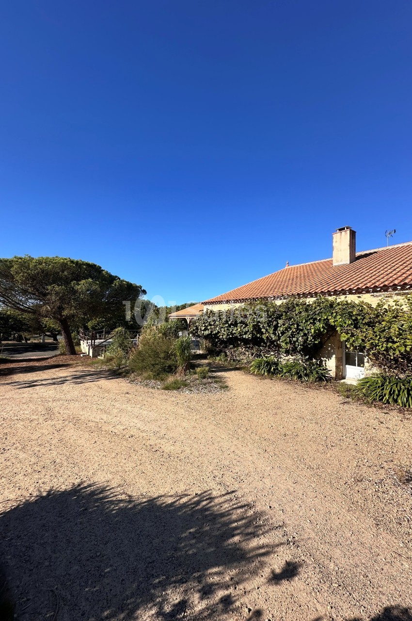 Bâtisse provençale en pierre avec toit de tuiles, entourée de végétation méditerranéenne et d'une cour en graviers.