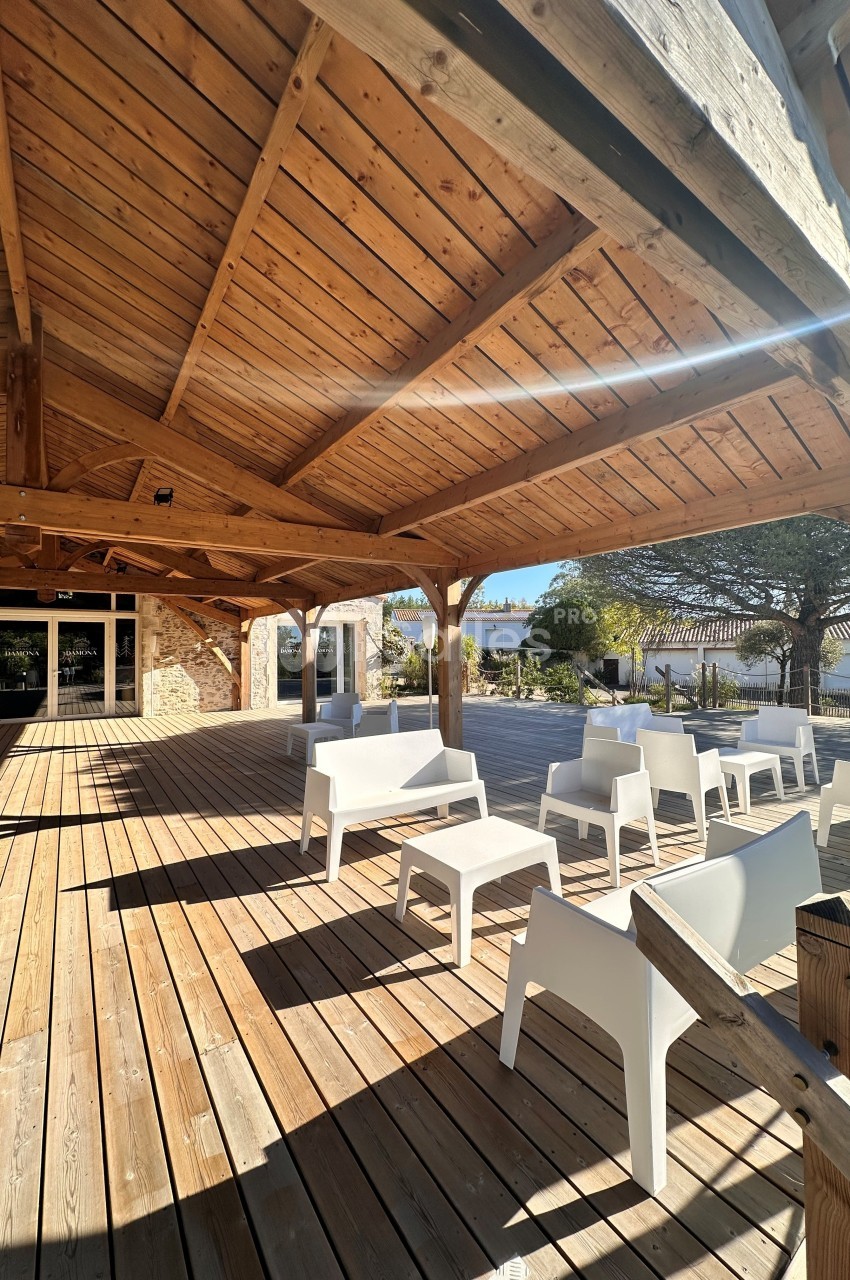 Terrasse couverte avec charpente bois, sol en lames bois clair, chaises blanches et bâtiments de pierre en arrière-plan.