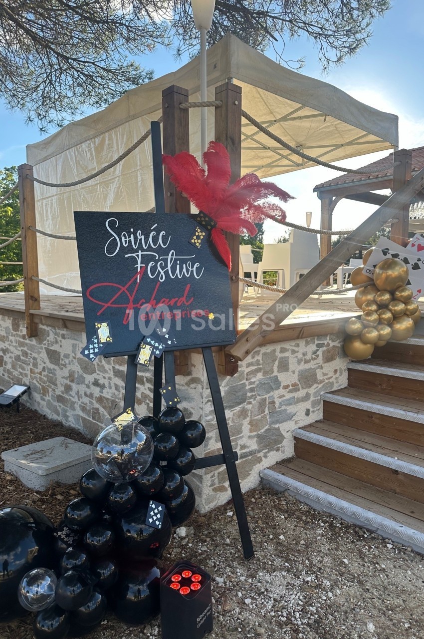Terrasse extérieure décorée avec panneau événementiel, ballons noirs et ornements rouges et dorés.