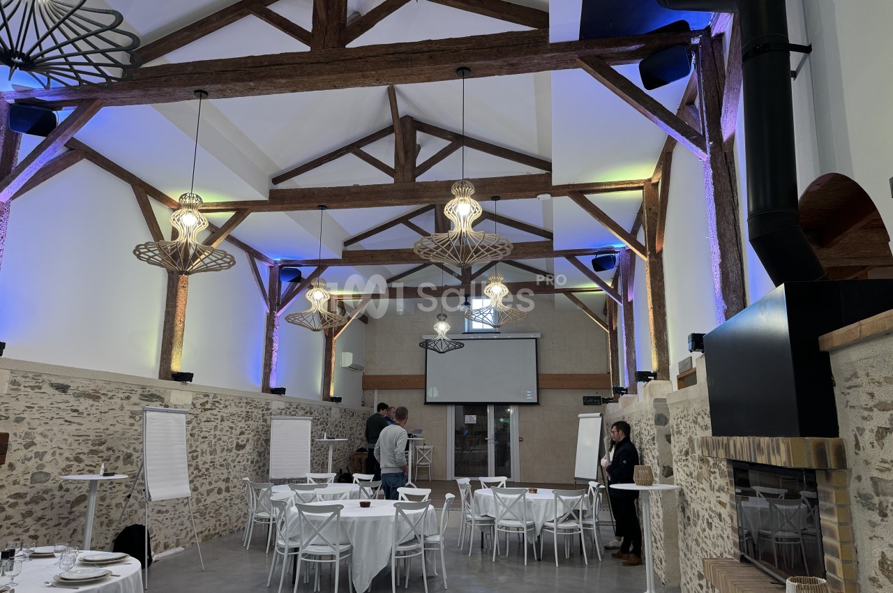Salle intérieure avec charpente apparente en bois, murs en pierre, éclairage bleu ambiant et tables dressées pour réception.