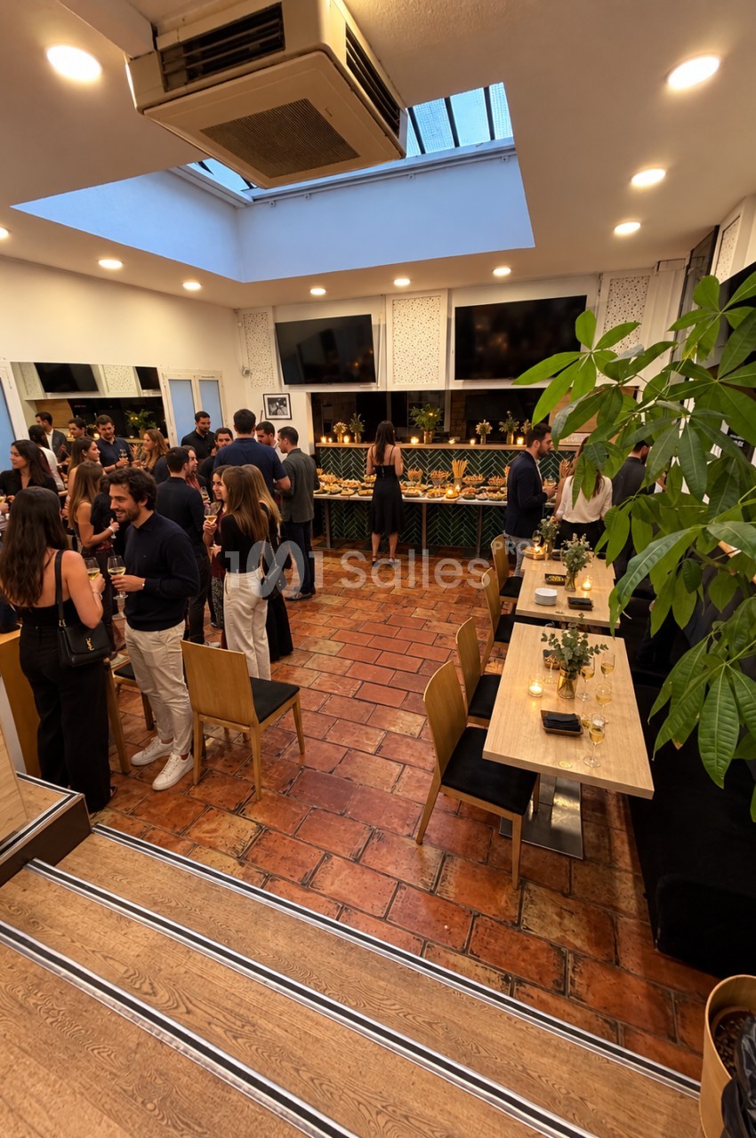 Salle intérieure avec convives debout et attablés, sol en tomettes, verrière zénithale, tables dressées et plantes vertes.
