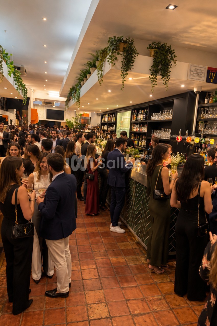 Salle intérieure de cocktail avec environ 150 convives, bar en arrière-plan, plantes suspendues, éclairage tamisé et sol en carrelage terracotta.