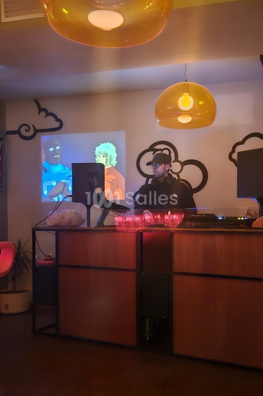 DJ en cabine derrière un pupitre bois, équipement audio visible, écrans d'ambiance colorés, luminaires suspendus dorés, décoration nuages stylisés.