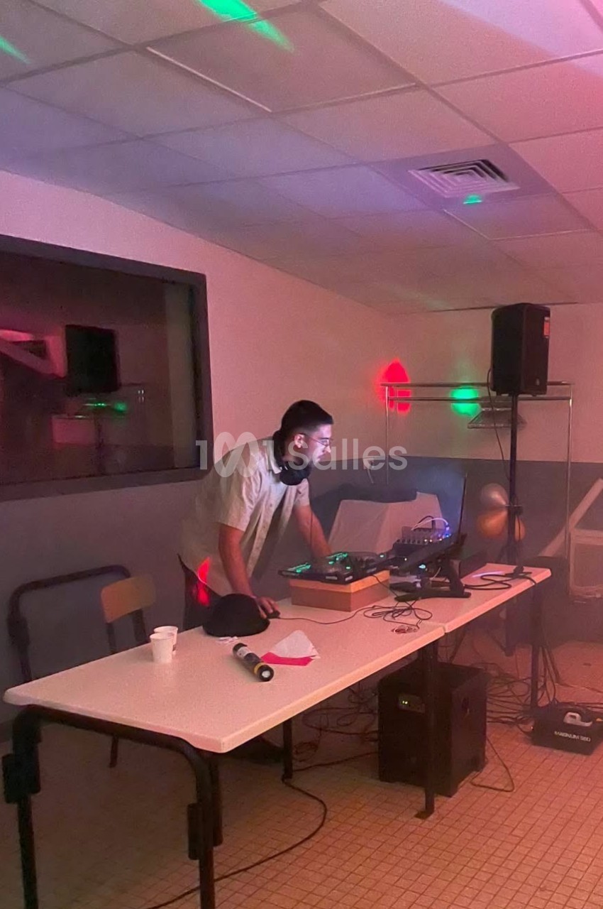 DJ en stand en intérieur face à sa console, éclairage rose et vert, enceintes et équipement autour.
