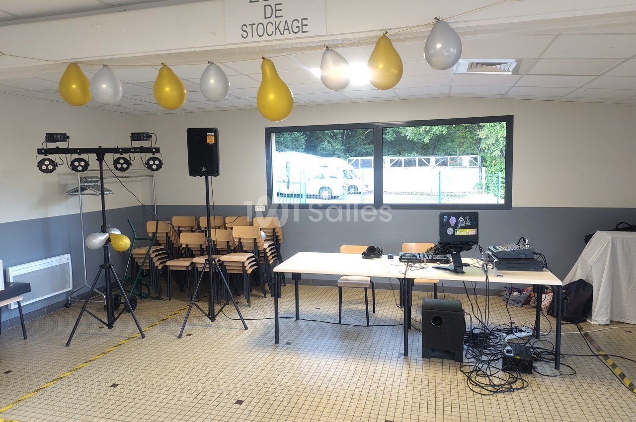 Salle intérieure équipée de projecteurs, matériel DJ sur table, chaises empilées et ballons jaunes et blancs accrochés au plafond.