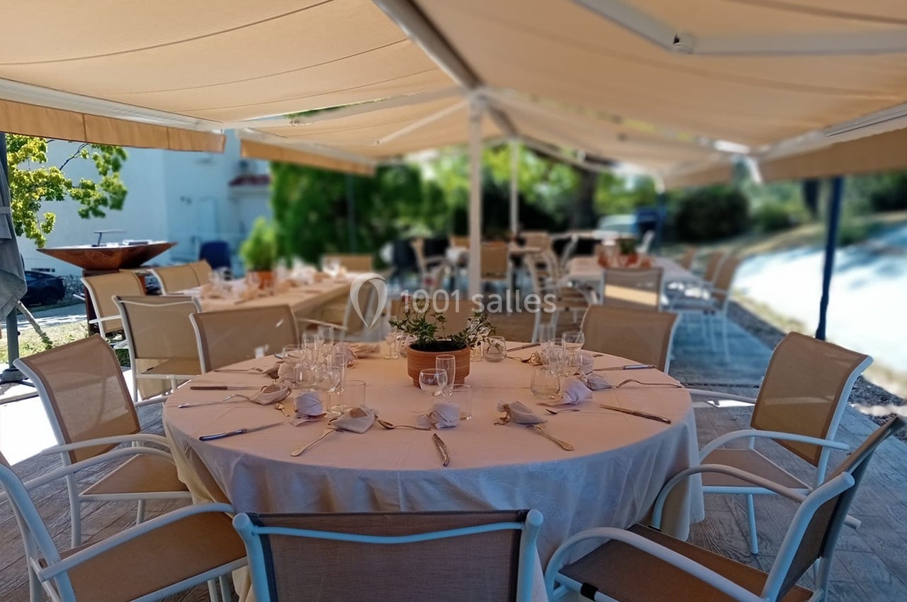 Table ronde dressée avec vaisselle blanche sous une terrasse ombragée, entourée de chaises et de verdure.