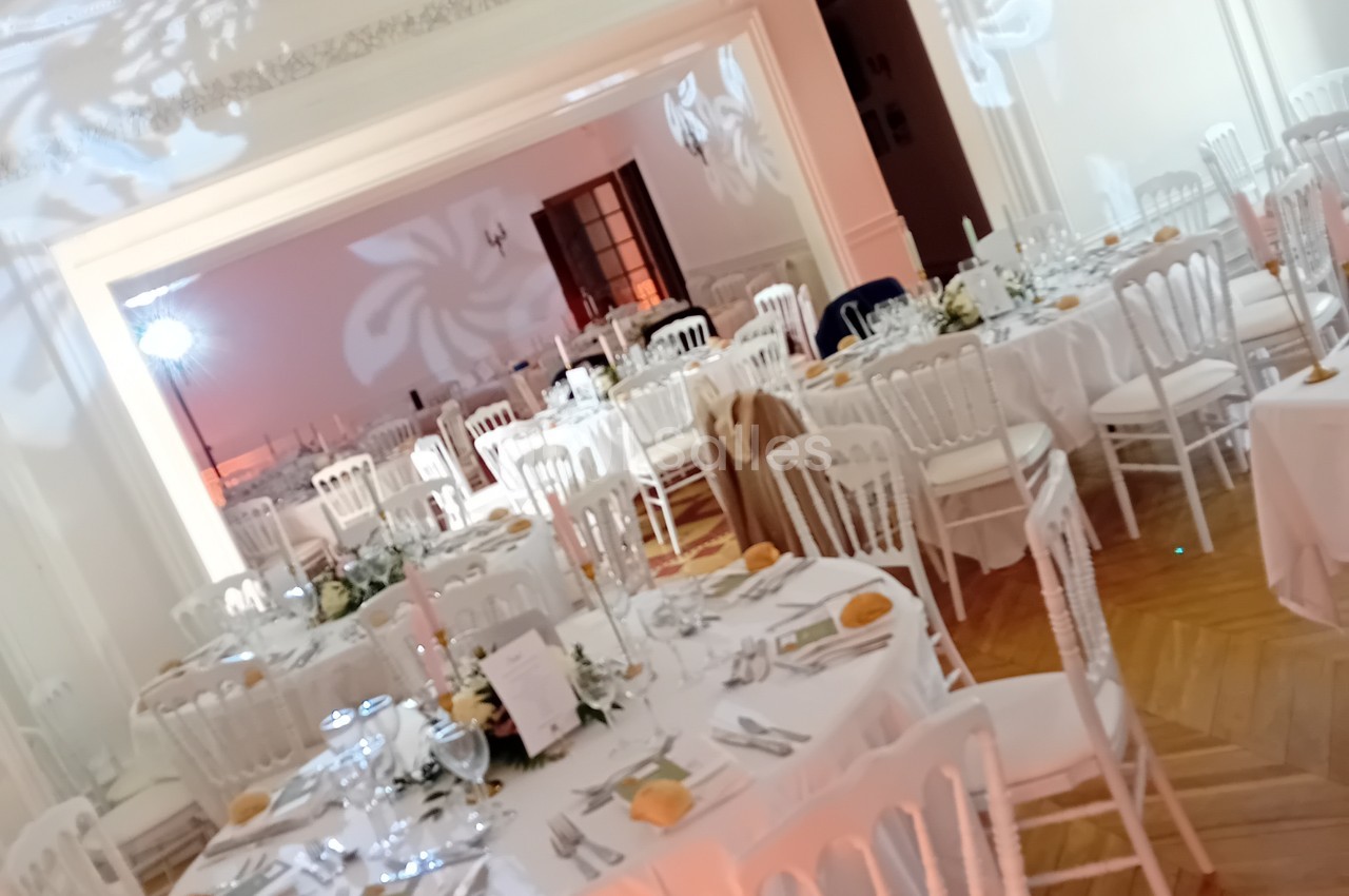 Salle dressée pour réception avec tables rondes nappe blanche, chaises chiavari blanches et noires, décoration florale et éclairage rose ambiant.