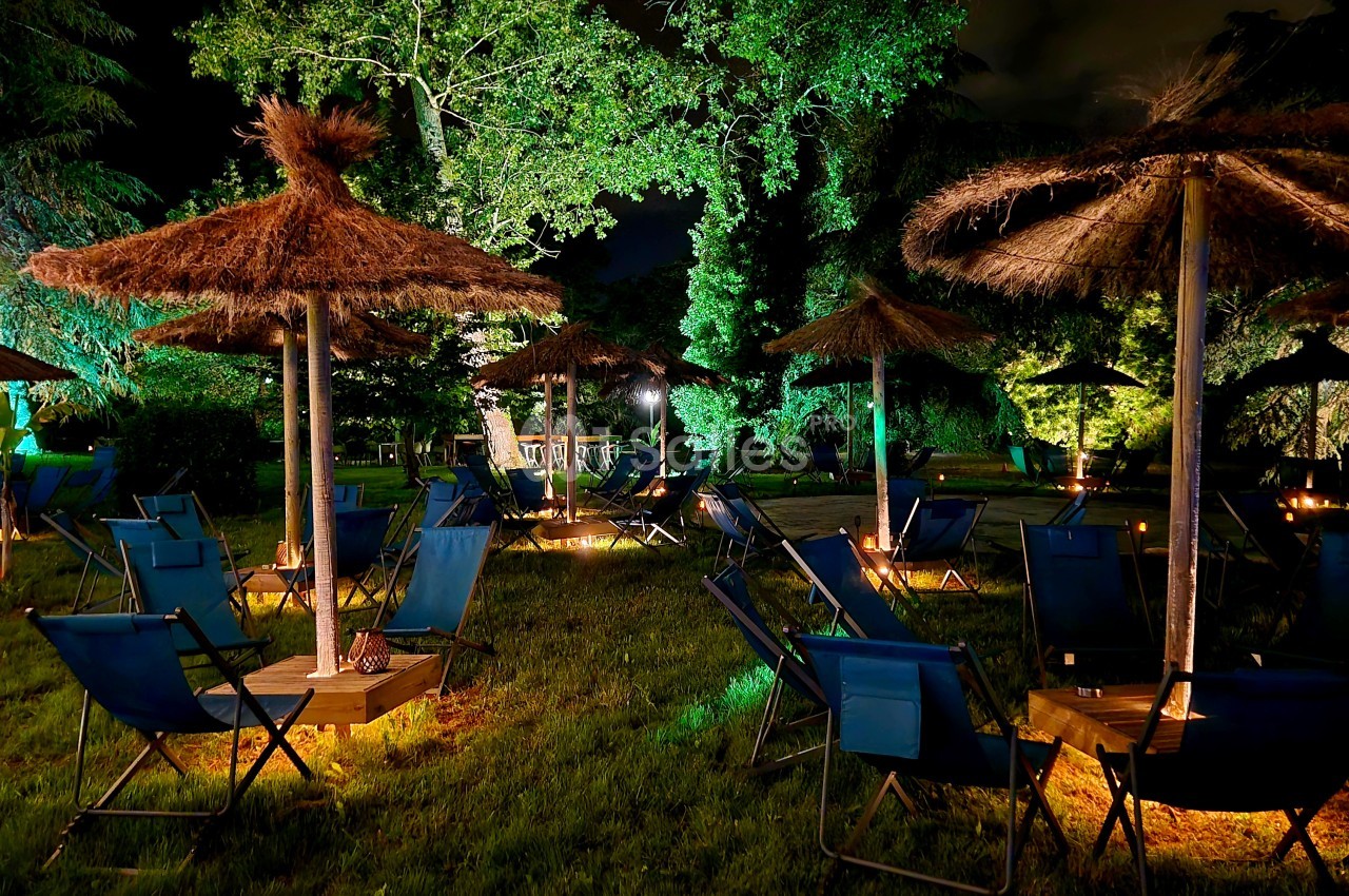 Espace extérieur de nuit avec transats bleus dispersés sous des parasols en paille, arbres illuminés en vert, pelouse et éclairage chaleureux au sol.