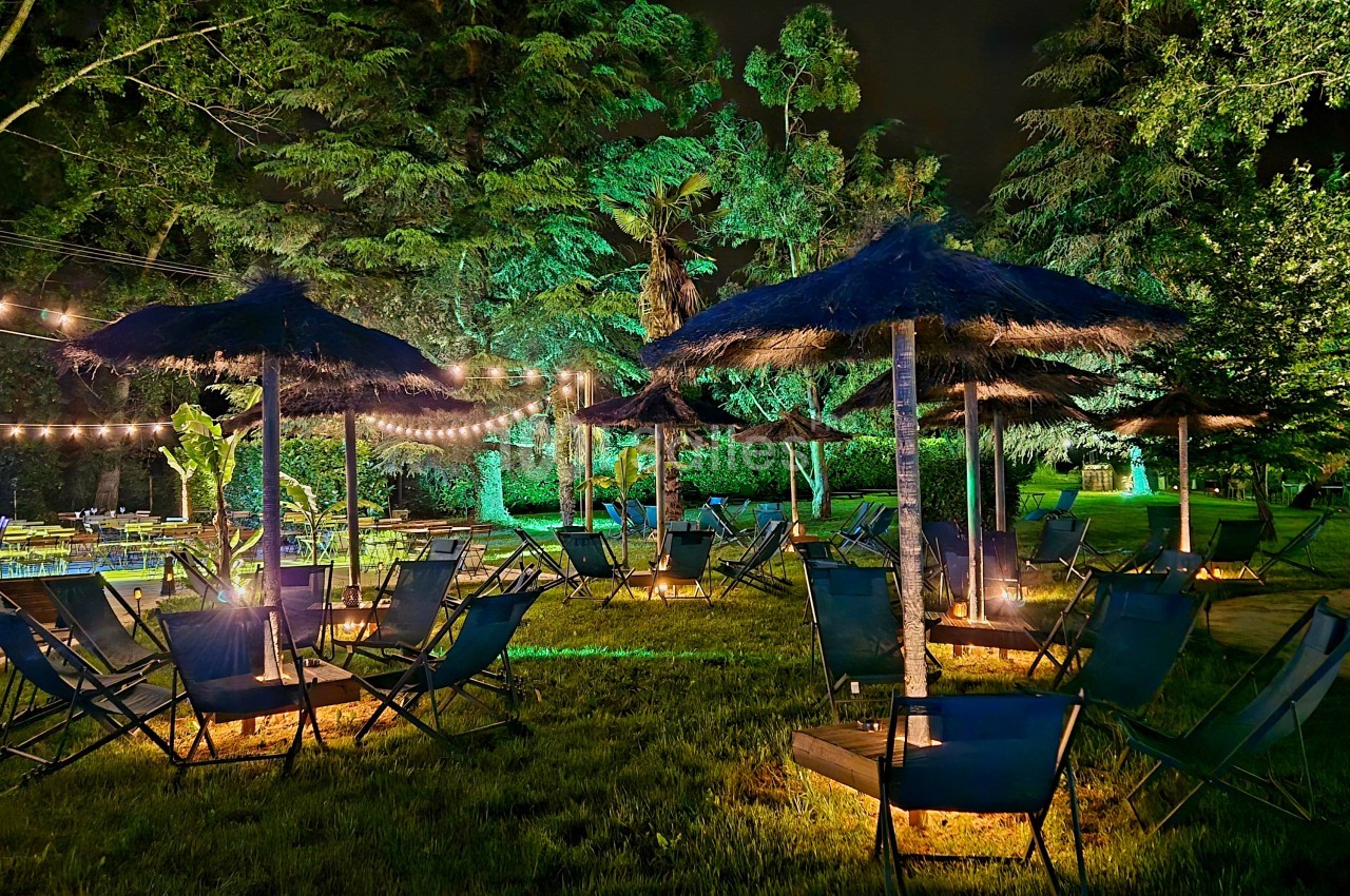 Jardin de nuit avec chaises longues sous des parasols en paille et éclairage ambiant - arbres verdoyants en arrière-plan.