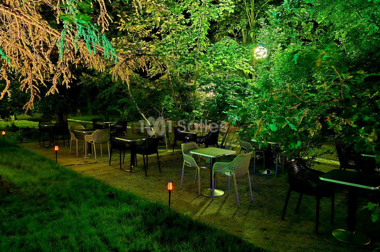 Terrasse de jardin en soirée avec tables et chaises dressées sous les arbres, éclairage des lanternes.