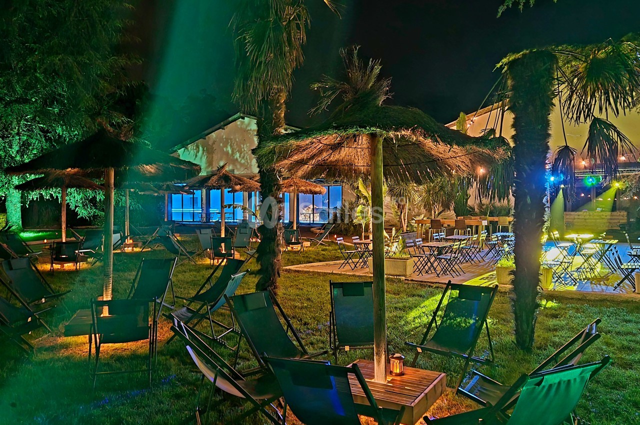 Espace extérieur en soirée avec chaises longues et parasols en paille, palmiers illuminés, vue sur un bâtiment en retrait.