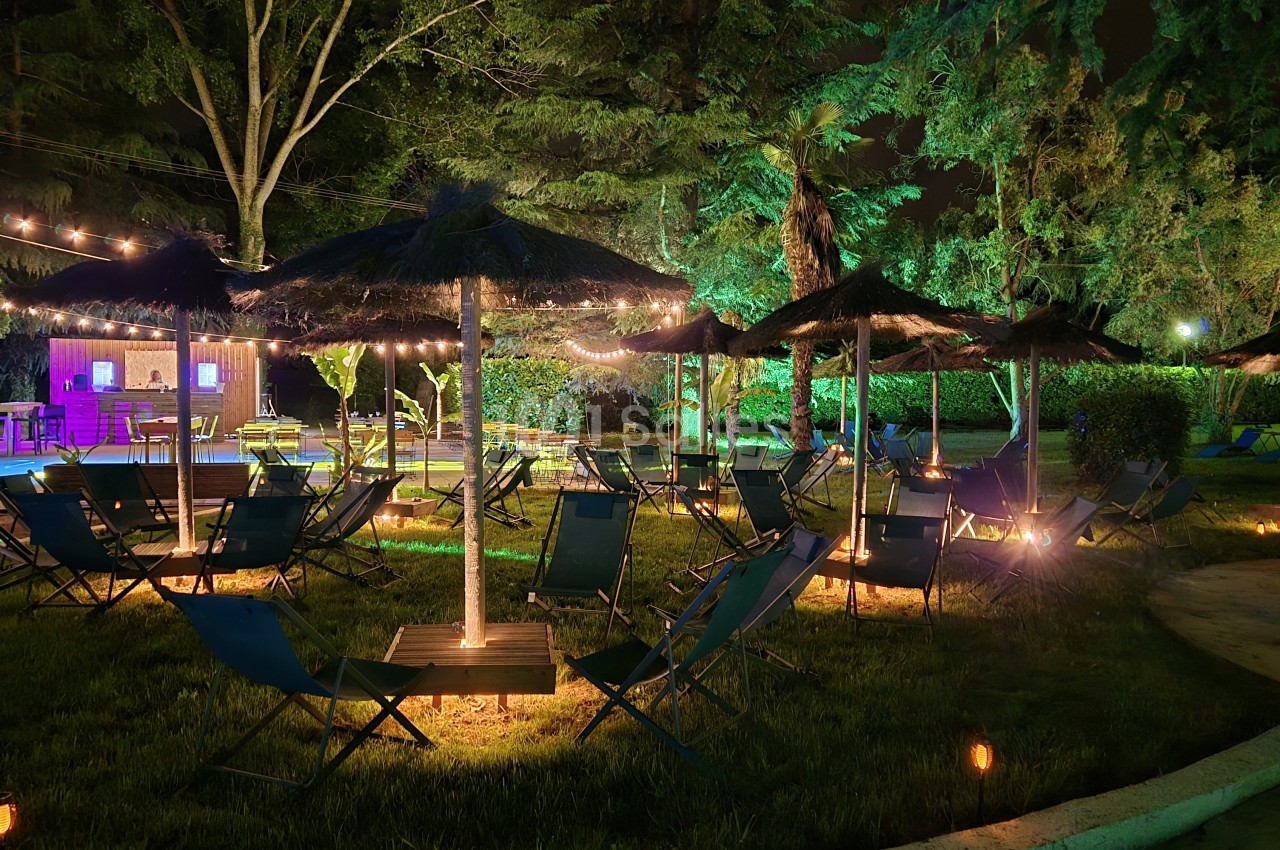 Espace extérieur de nuit avec chaises longues sous parasols, guirlandes lumineuses et arbres éclairés au-dessus d'une pelouse.