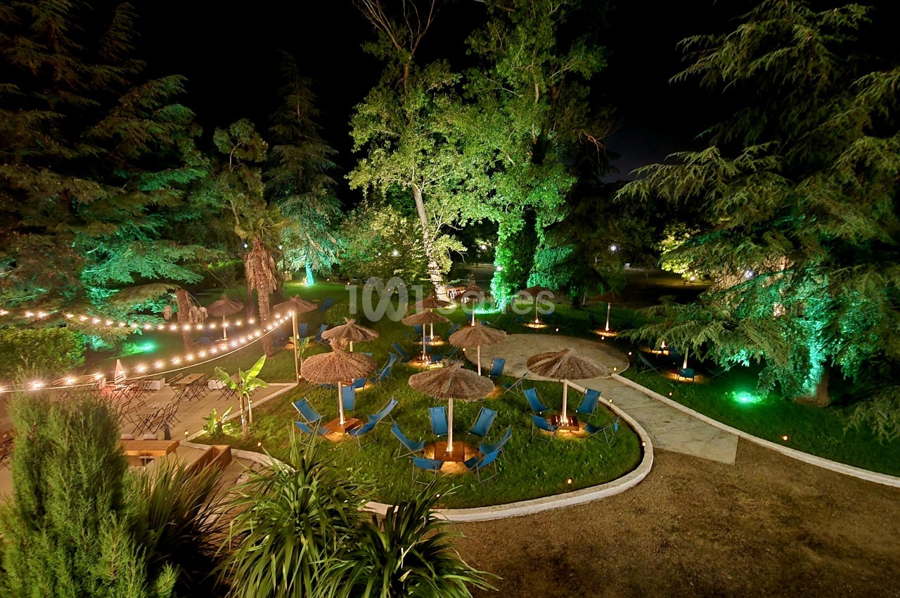 Espace extérieur de nuit avec arbres éclairés en vert, chaises longues bleues sous parasols et guirlandes lumineuses.