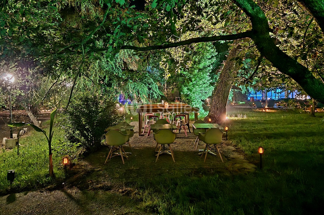 Espace extérieur de nuit sous les arbres avec tables dressées, chaises vertes et éclairage tamisé au sol.