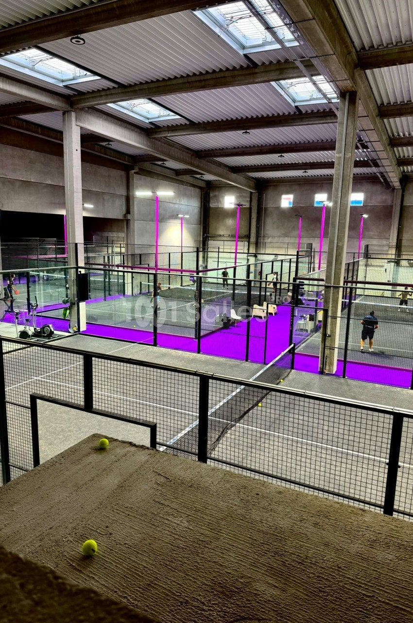 Salle intérieure avec plusieurs terrains de padel entourés de grillages, éclairée par des néons violets et des fenêtres en…