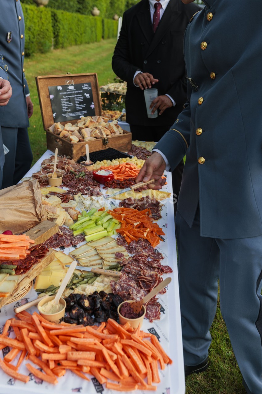 Plateau de charcuterie, crudités et canapés en plein air face à des convives en tenue formelle.