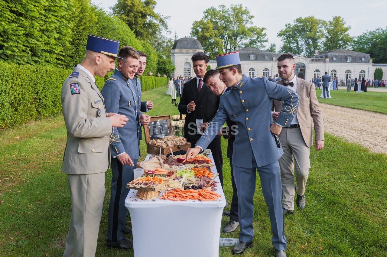 Militaires en uniforme autour d'une table de buffet froid en plein air, parc avec château en arrière-plan.