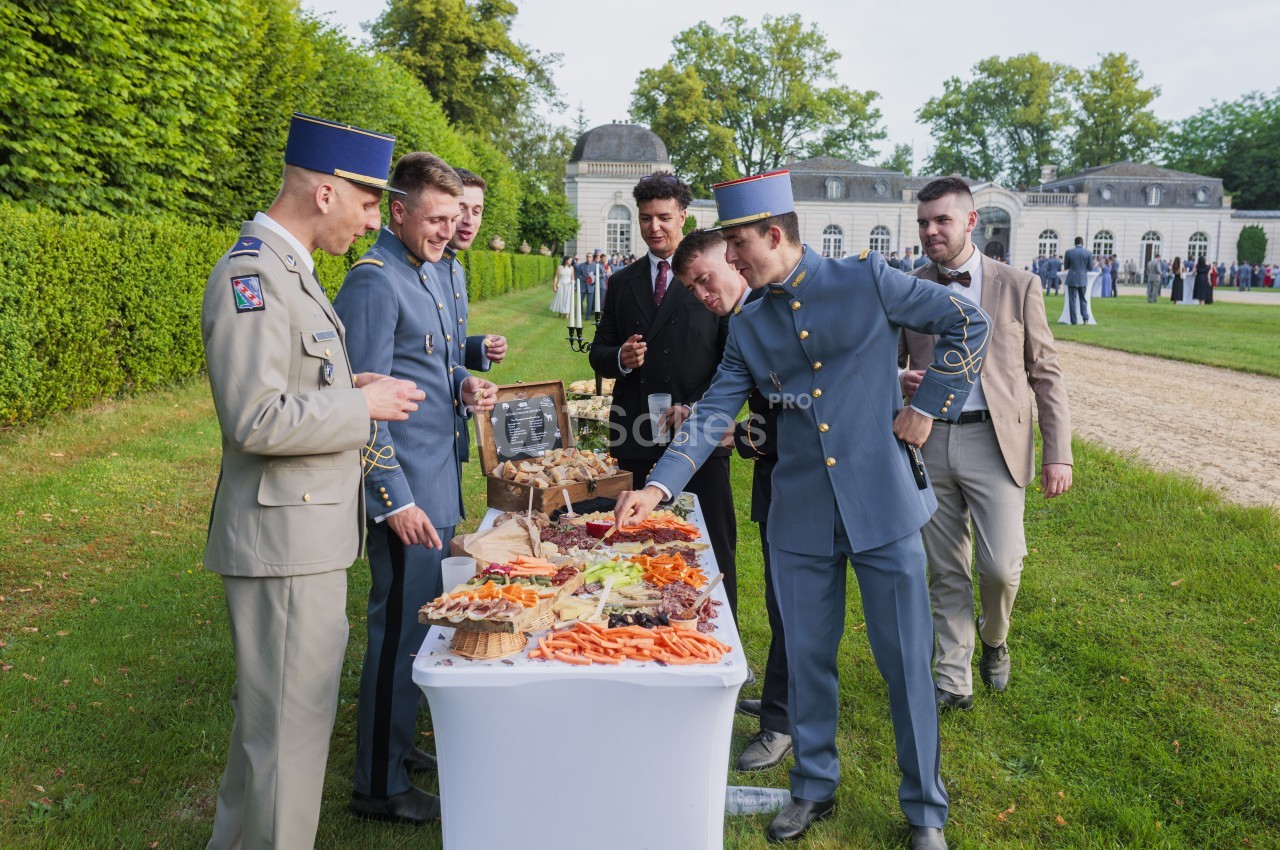 Militaires en uniforme autour d'une table de buffet froid en plein air, parc avec château en arrière-plan.