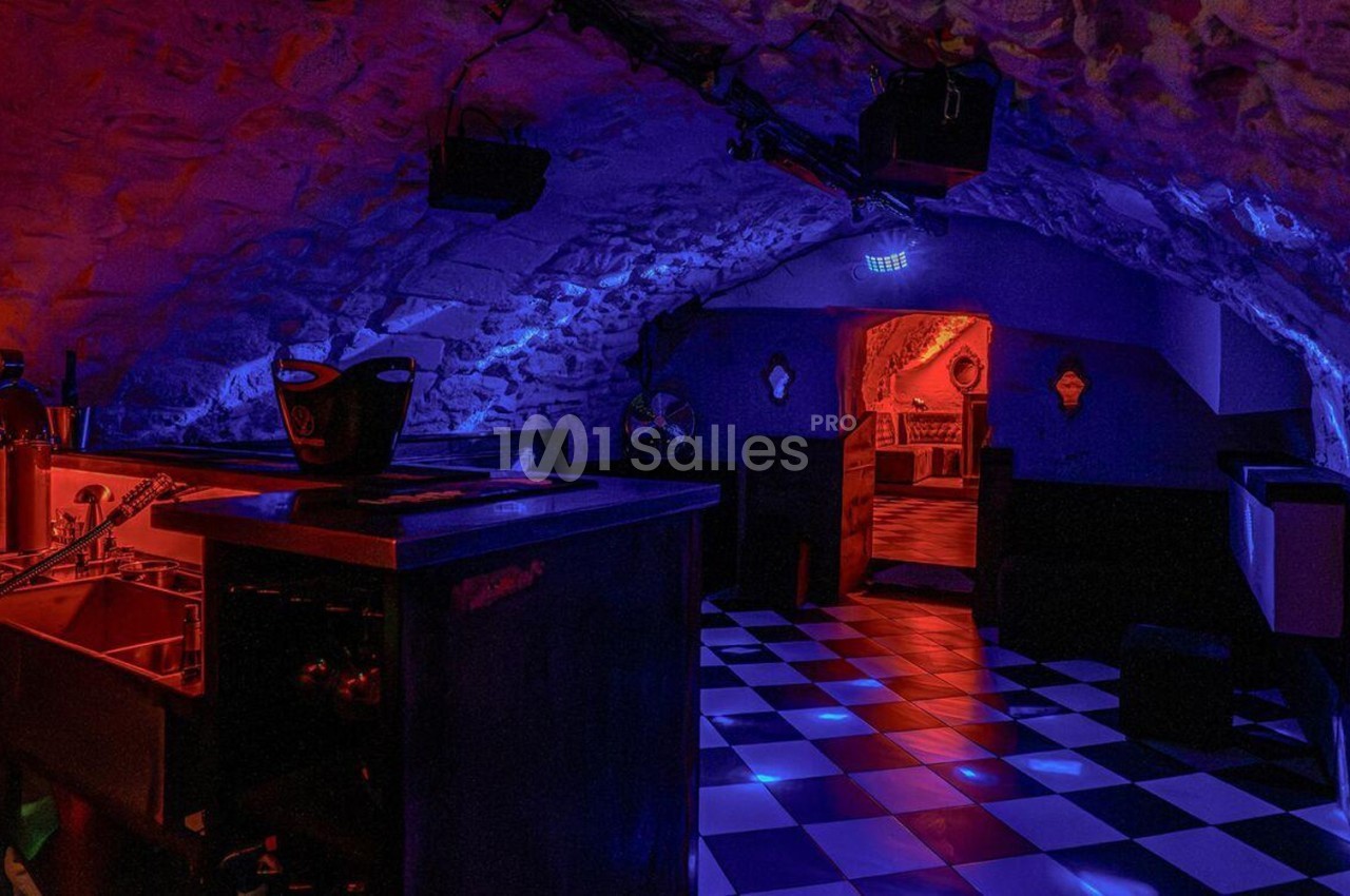 Salle voûtée avec éclairage tamisé rouge et bleu, sol en damier noir et blanc, bar et mobilier sombre.