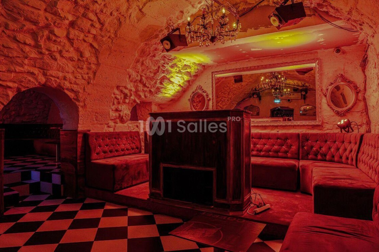 Salle voûtée en pierre avec banquettes rouges, mobilier en bois et sol à carreaux noirs et blancs.
