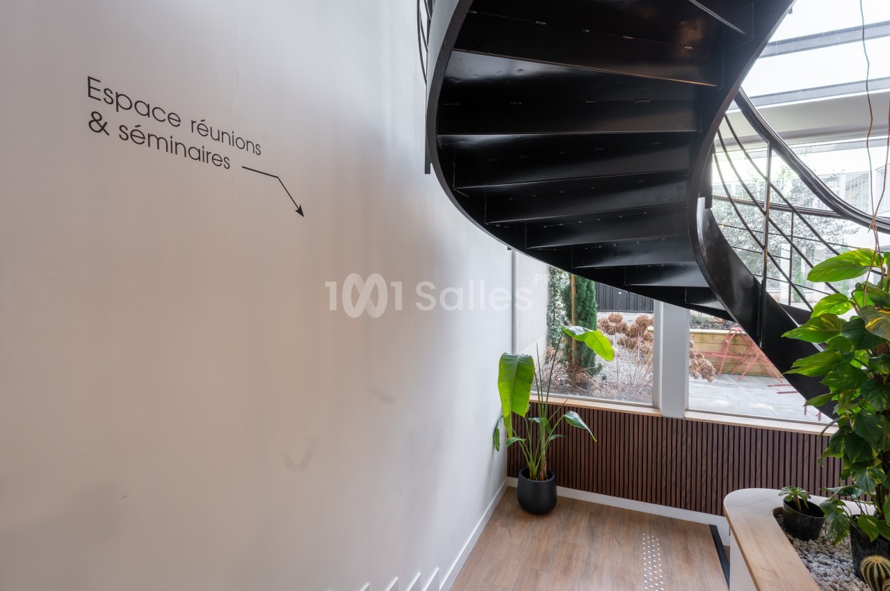 Escalier en spirale noir avec signalétique murale, plantes vertes et vue sur jardin intérieur.