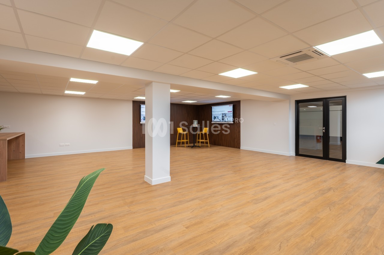 Salle spacieuse vide avec sol en bois clair, pilier blanc central et éclairage encastré aux plafonds.