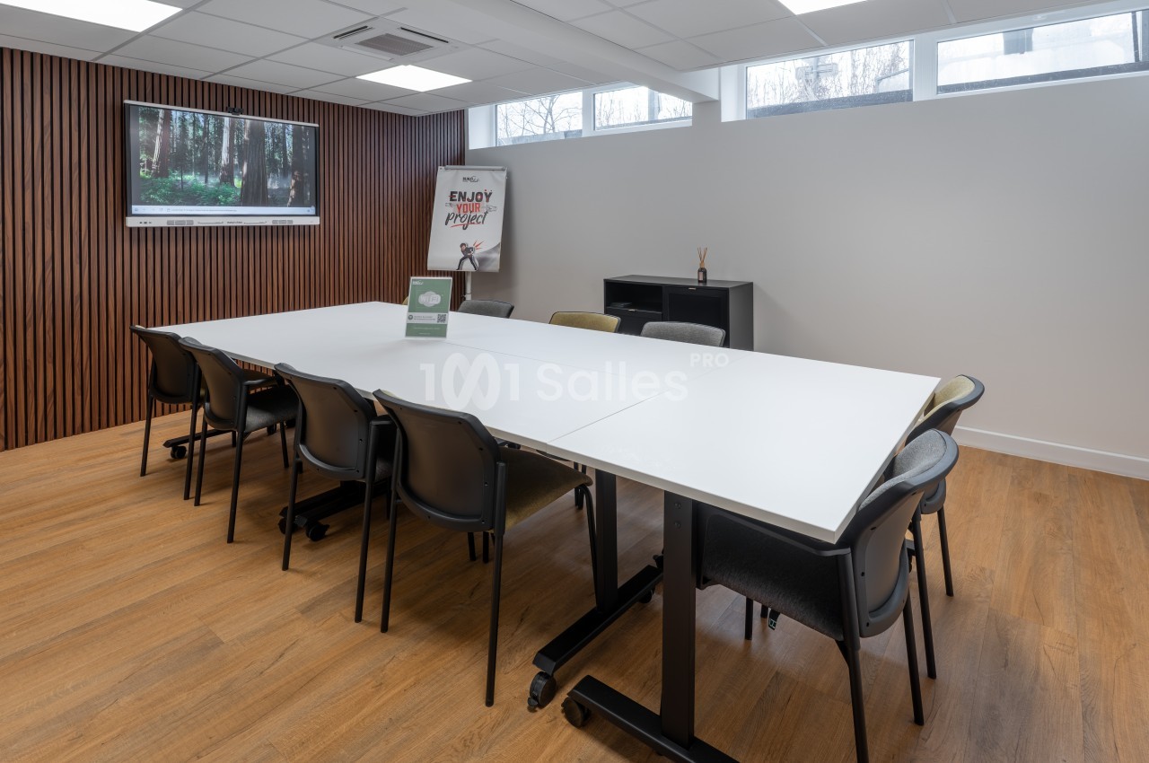 Salle de réunion avec table blanche rectangulaire, chaises noires, paroi en bois, écran mural et sol parquet.