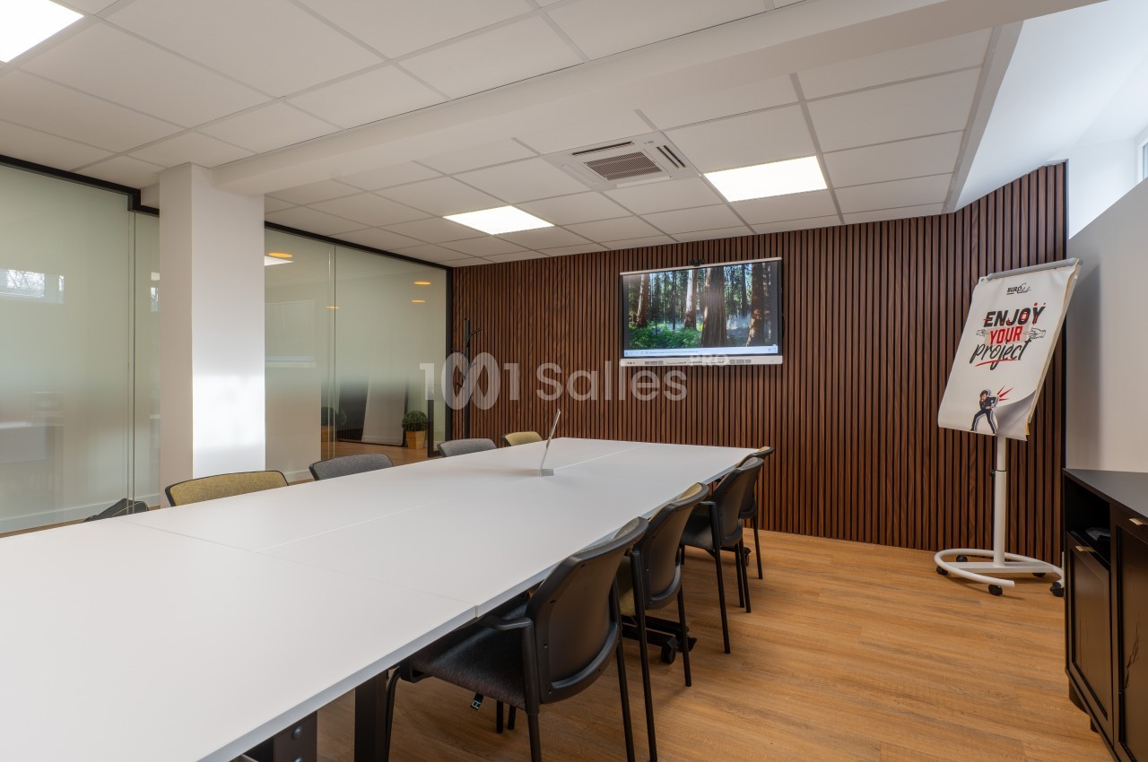 Salle de réunion avec table blanche rectangulaire, chaises noires, paroi bois, écran mural et paroi vitrée.