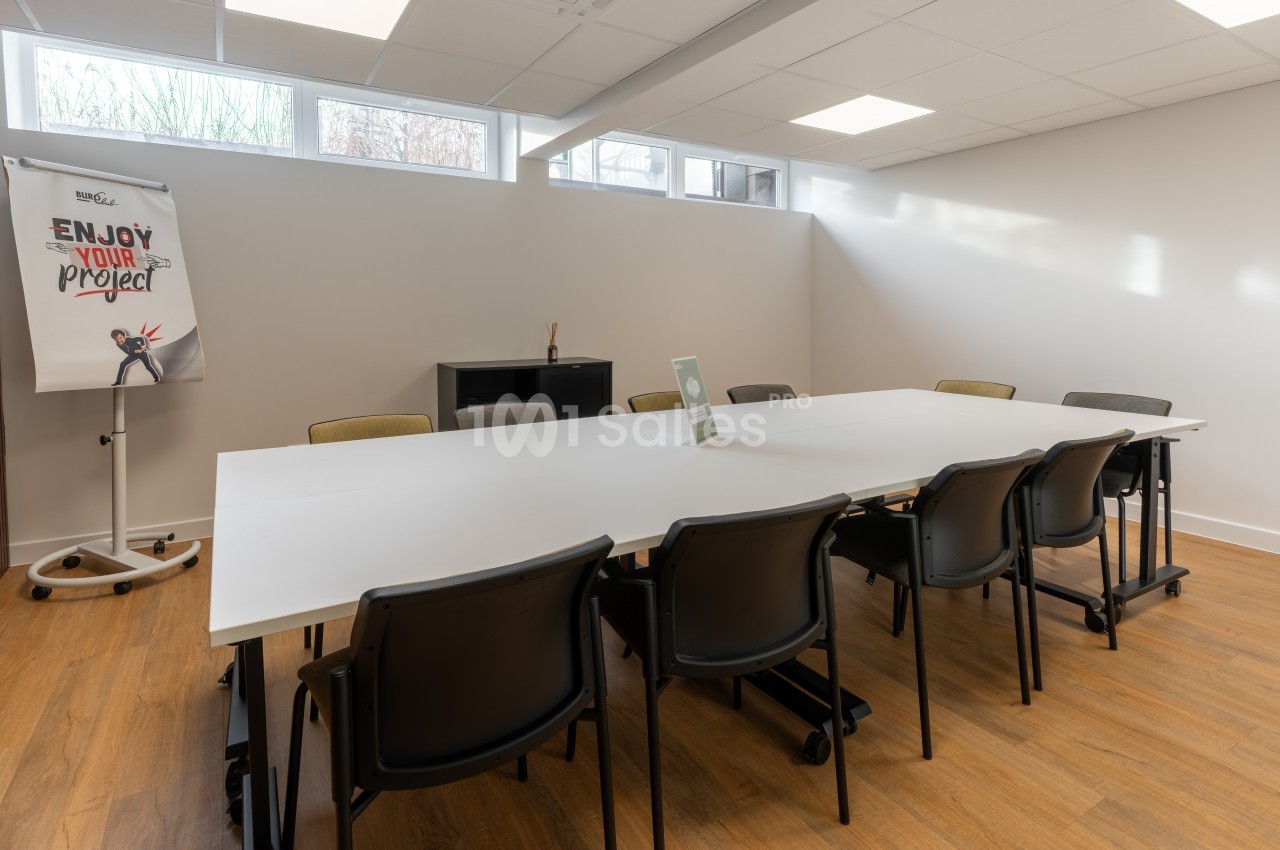 Salle de réunion avec table blanche rectangulaire, chaises noires, sol bois et éclairage naturel zénithal.