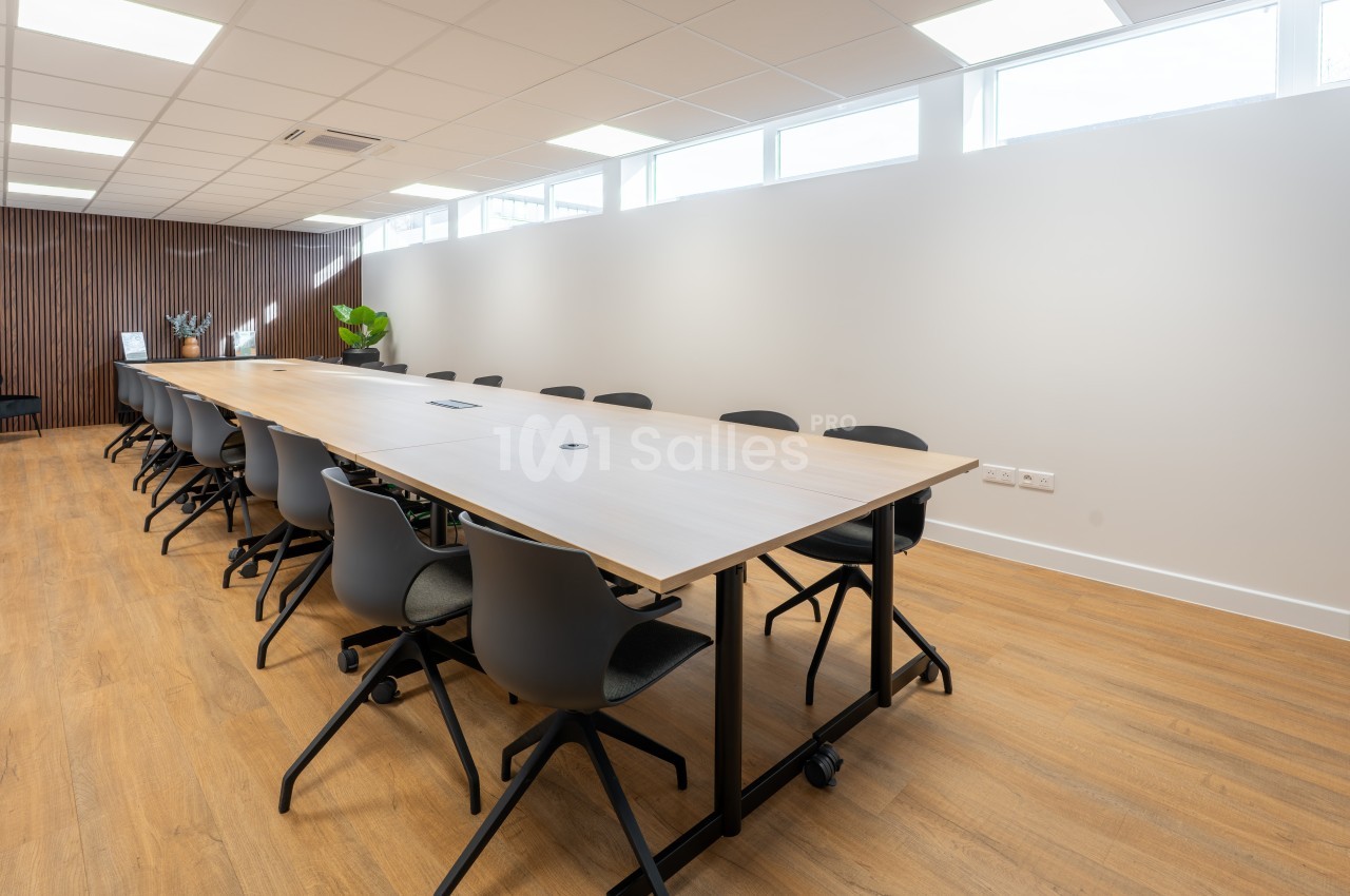 Salle de réunion avec table blanche rectangulaire, chaises noires et murs clairs - éclairage naturel zénithal.
