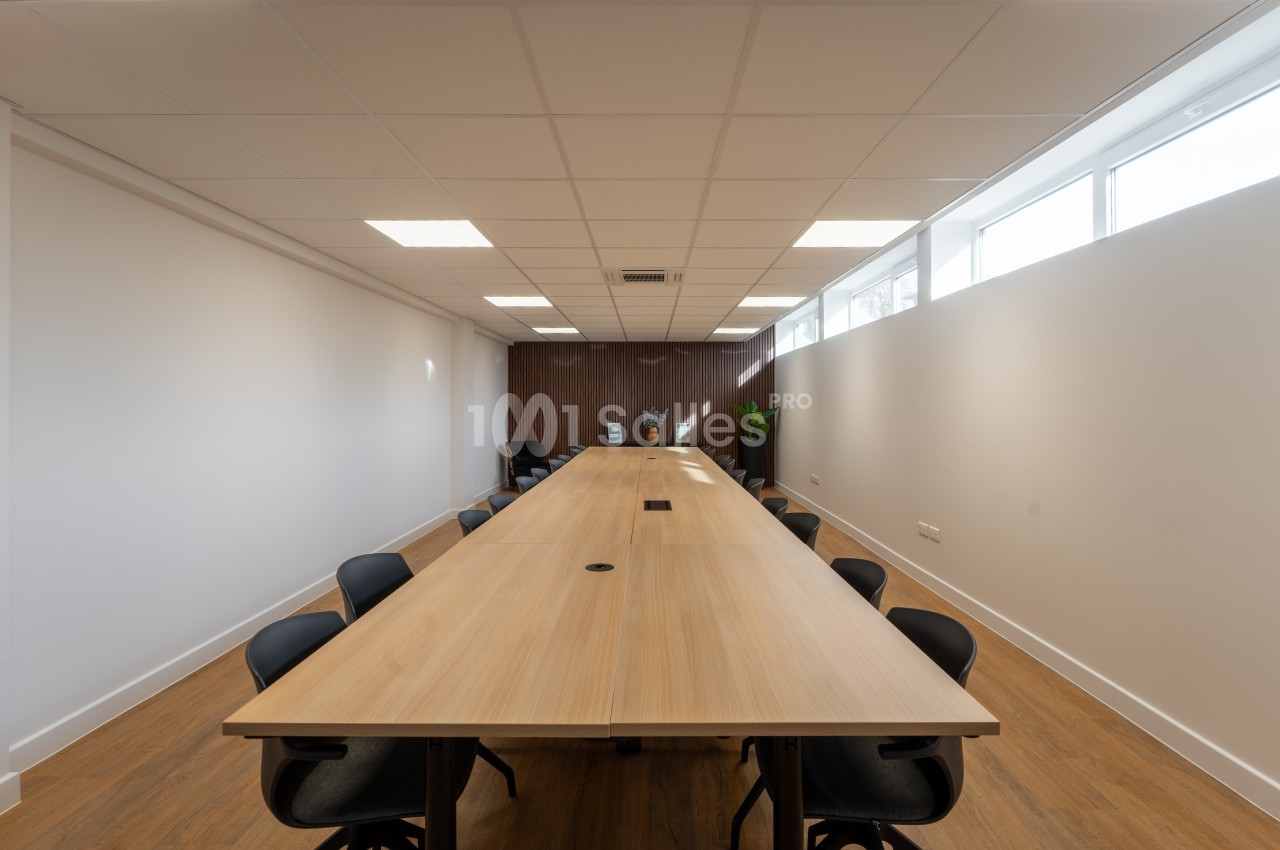 Salle de réunion avec table en bois clair et chaises noires - plafond suspendu et éclairage LED - baies vitrées.