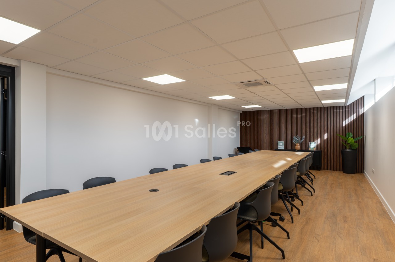 Salle de réunion avec table longue en bois, chaises noires et panneautage marron, éclairage plafond.