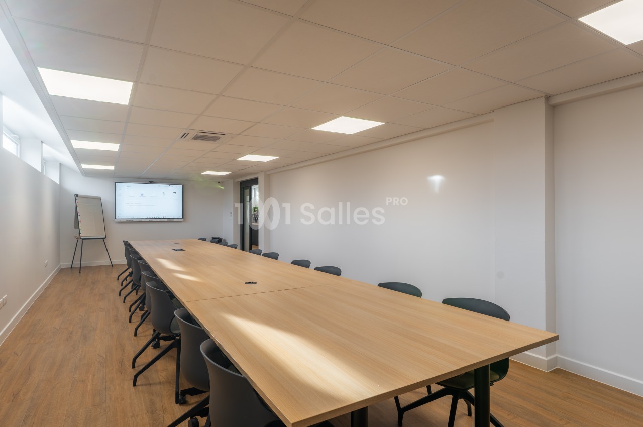 Salle de réunion avec table en bois, chaises noires, écran mural et éclairage encastré.