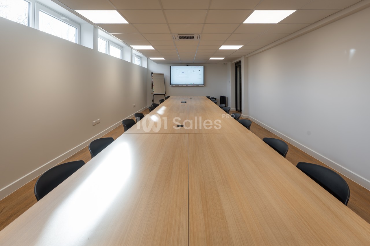 Salle de réunion avec table en bois clair, chaises noires et écran mural - éclairage zénithal.