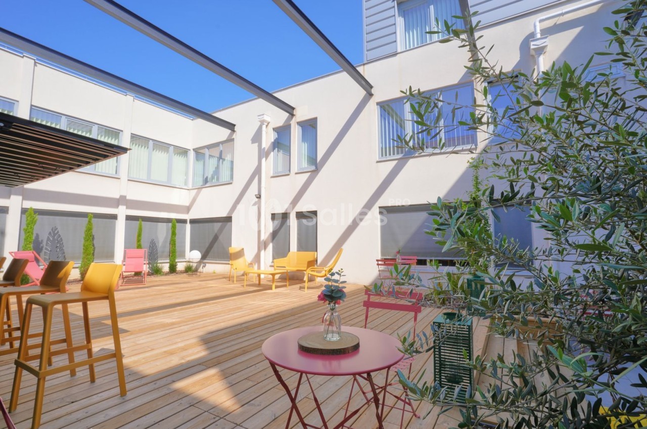 Terrasse ensoleillée avec mobilier coloré, tables basses et chaises hautes face à bâtiment blanc aux baies vitrées.