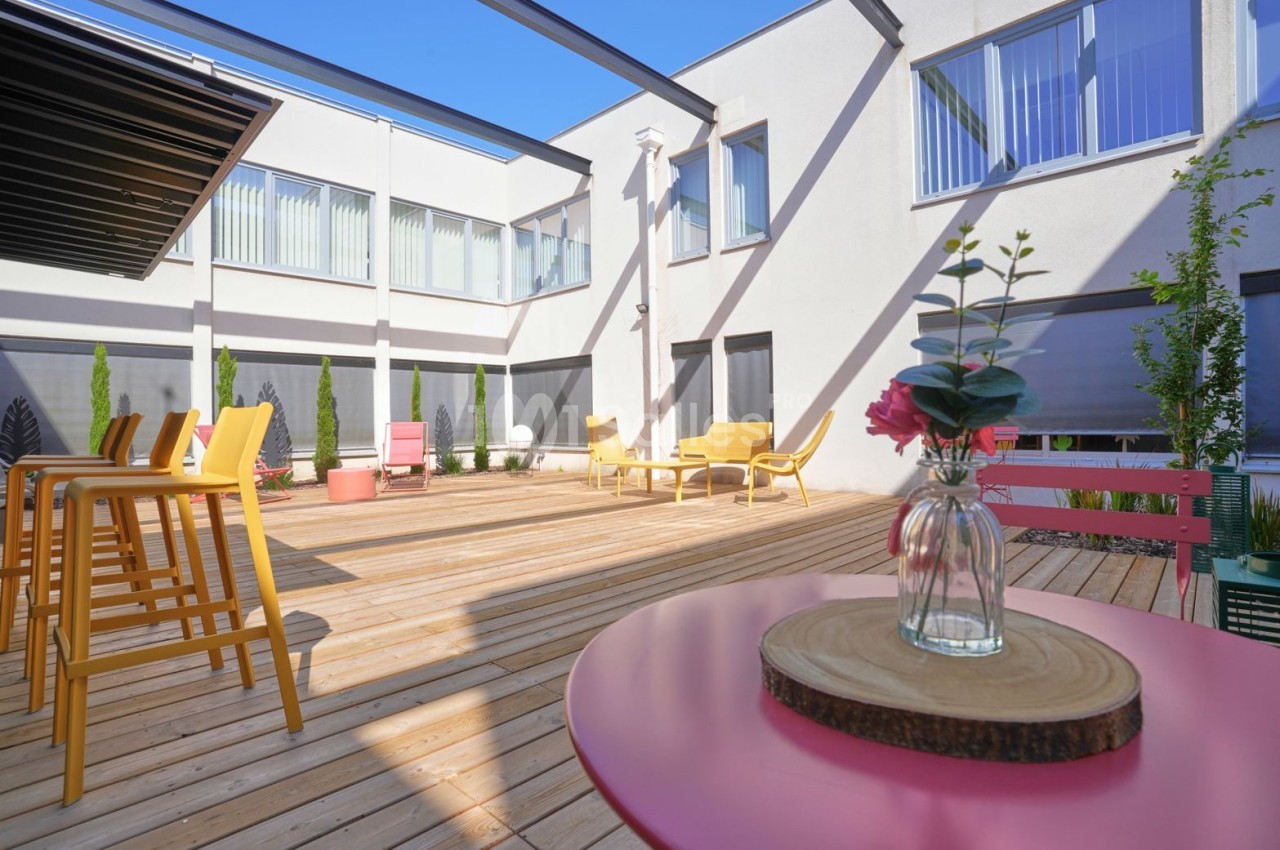 Terrasse extérieure aménagée avec mobilier jaune, tables rondes roses, plantes en pots et bâtiment blanc aux baies vitrées.