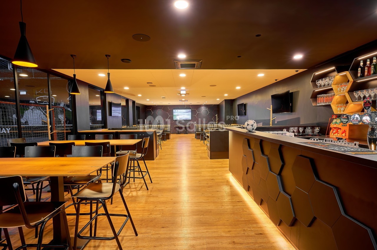 Intérieur de bar-lounge aménagé avec tables hautes en bois, comptoir, étagères de bouteilles et écrans TV - ambiance chaleureuse.