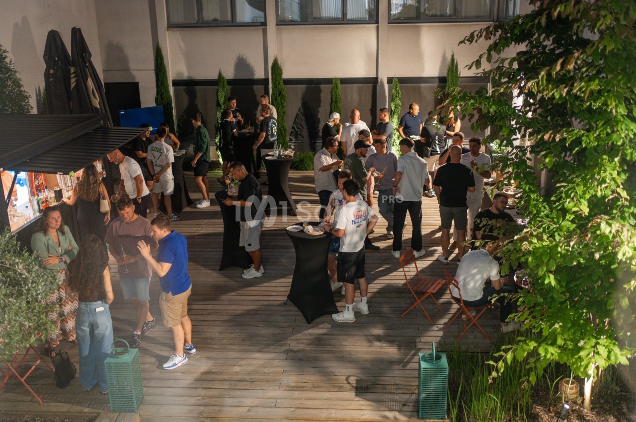 Terrasse en bois avec convives debout et assis, tables hautes noires, plantes vertes, façade vitrée en arrière-plan.