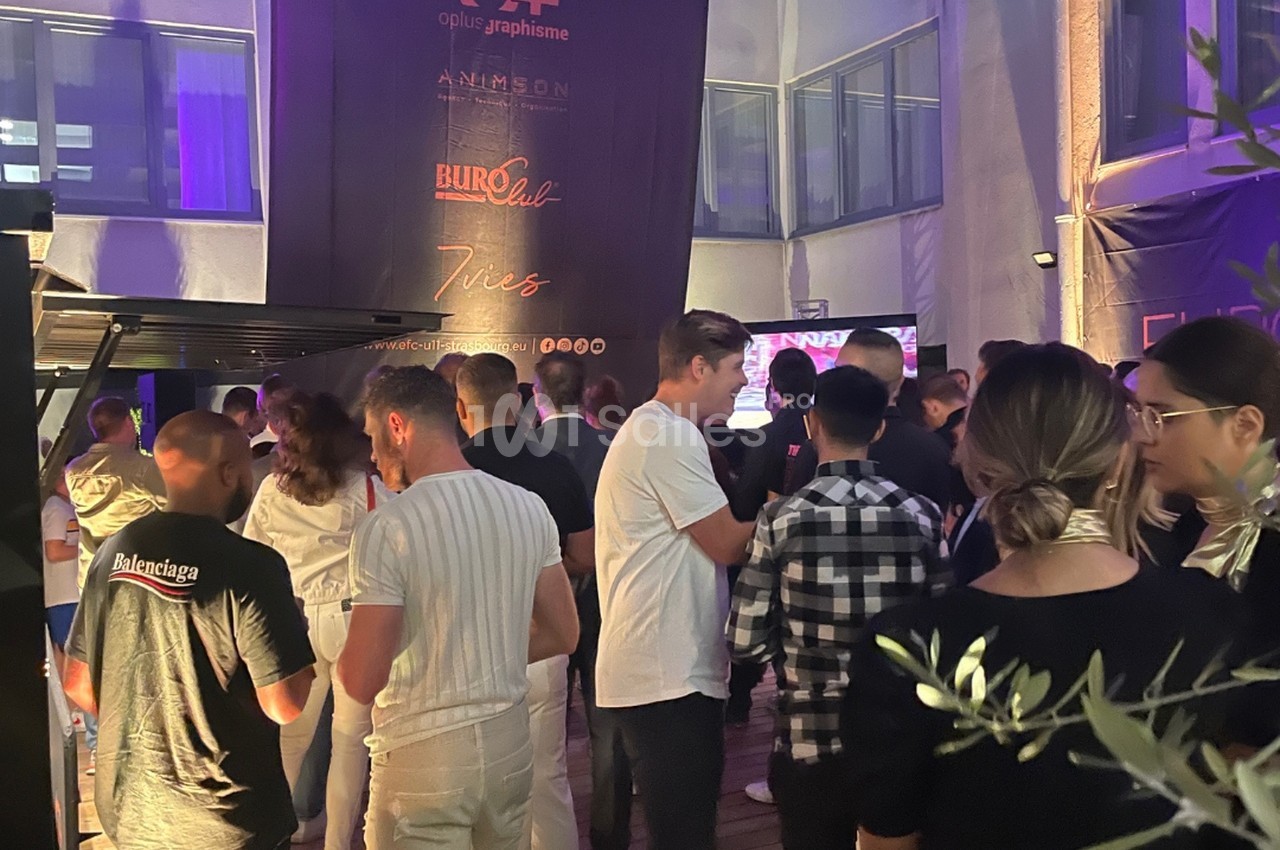 Salle de réception bondée avec éclairage violet, convives debout discutant, signalétique murale illuminée en arrière-plan.