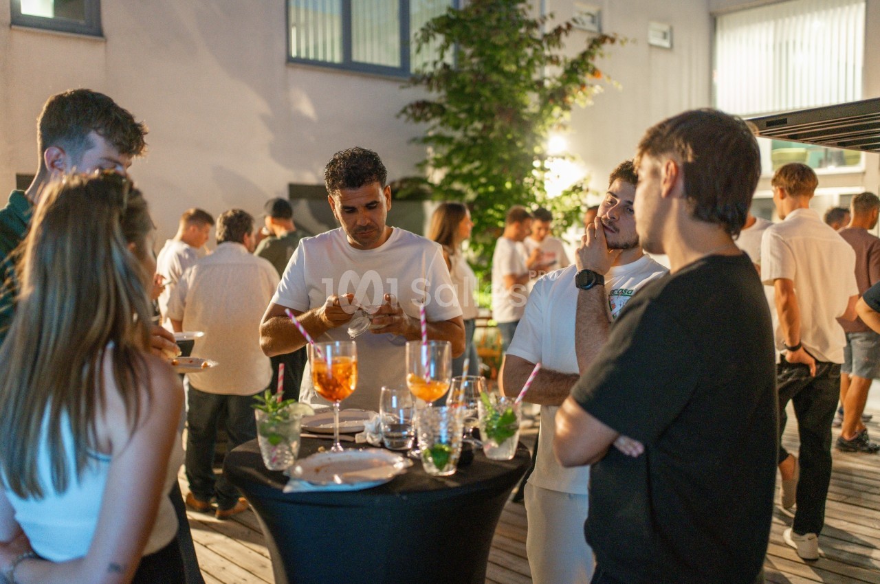 Espace intérieur lumineux avec convives debout autour de tables hautes, verres à la main, plantes vertes décoratives en arrière-plan.