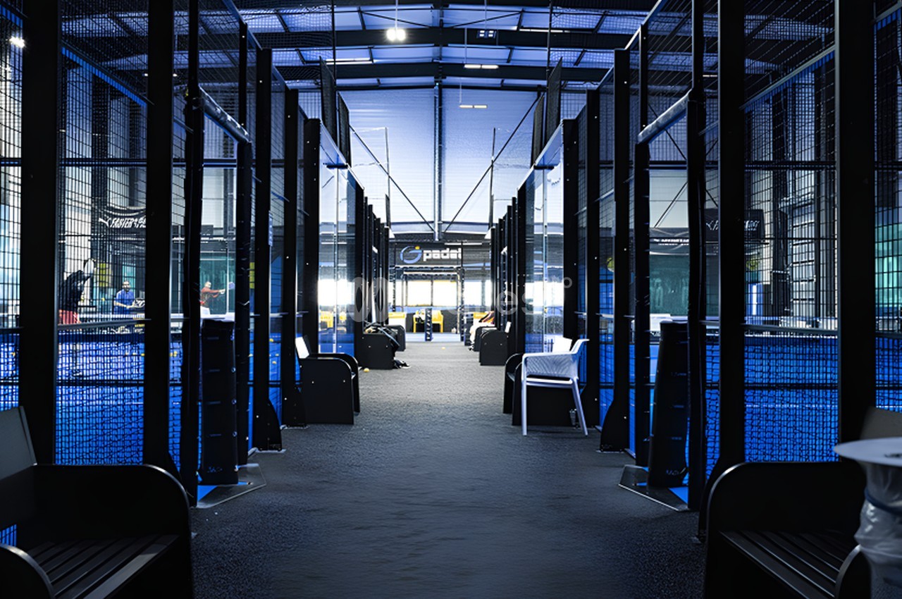 Couloir de terrain de padel couvert avec parois grillagées bleues et noires, accès aux courts à travers structure métallique.