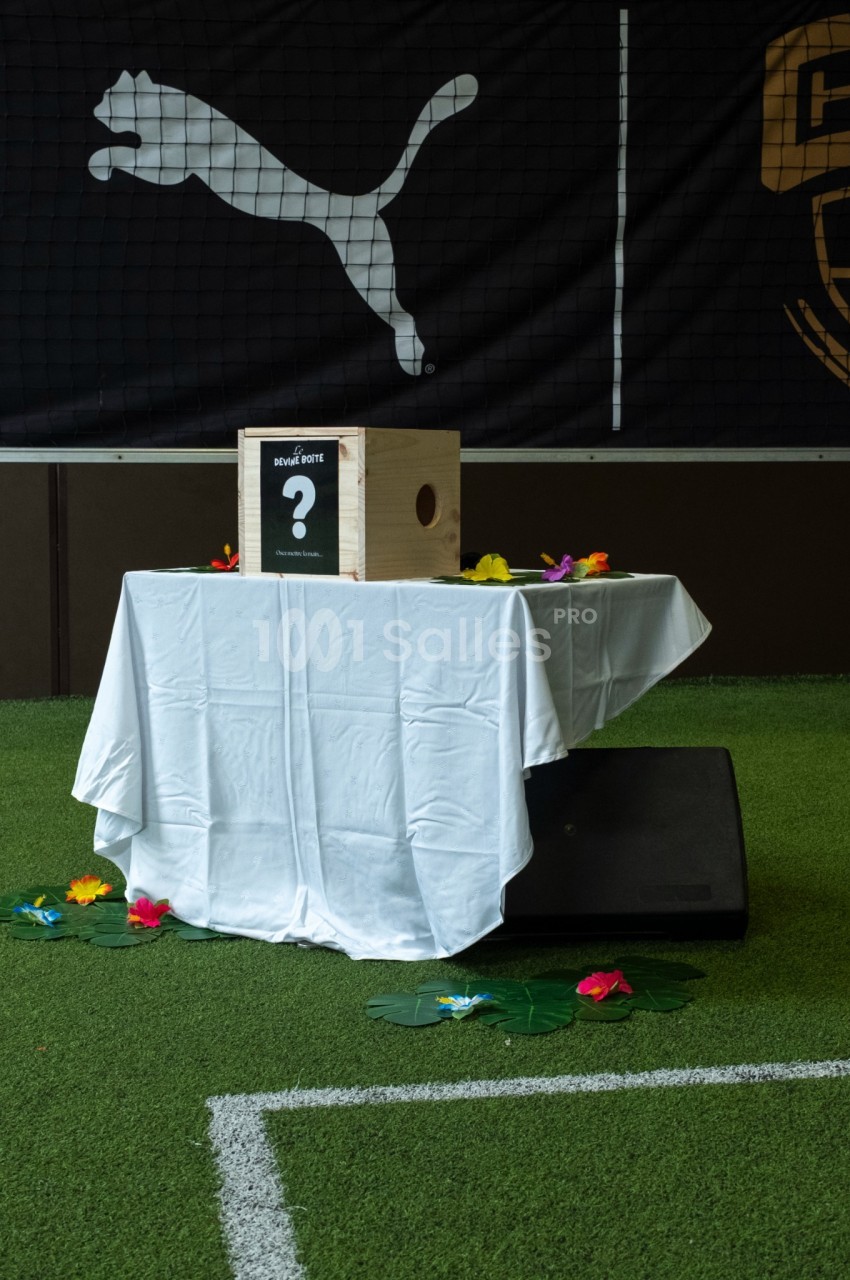 Table ronde dressée de blanc sous un logo Puma, décorée de fleurs colorées et d'une boîte en bois sur gazon synthétique.