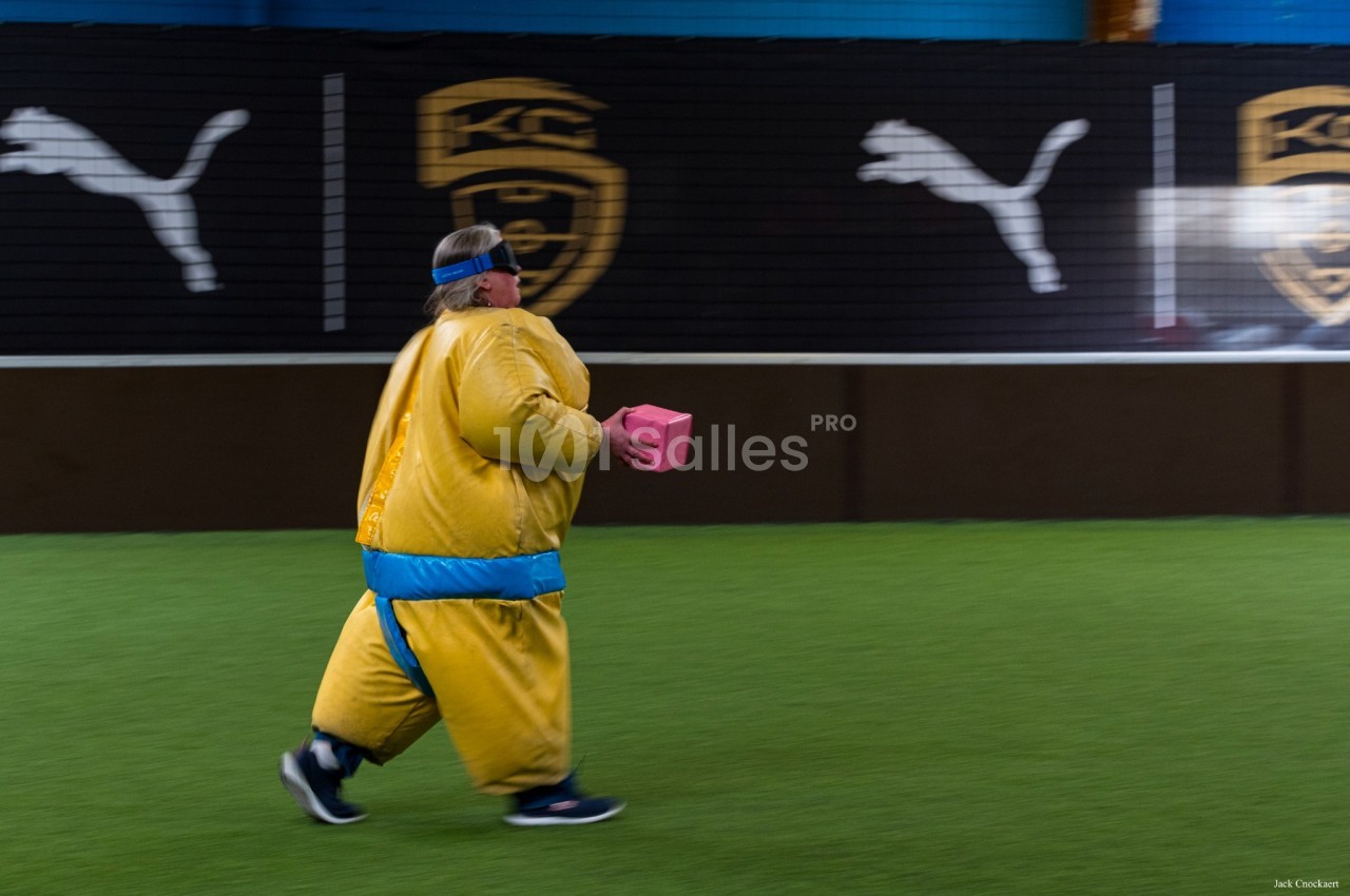 Personne en costume jaune et bleu courant sur terrain de baseball synthétique intérieur.