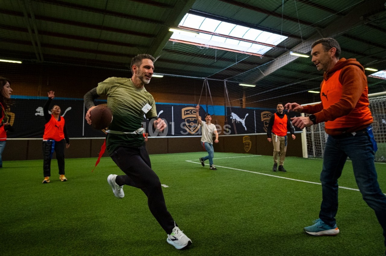 Terrain de football couvert avec gazon synthétique, groupe de personnes jouant au ballon américain sous éclairage naturel et artificiel.
