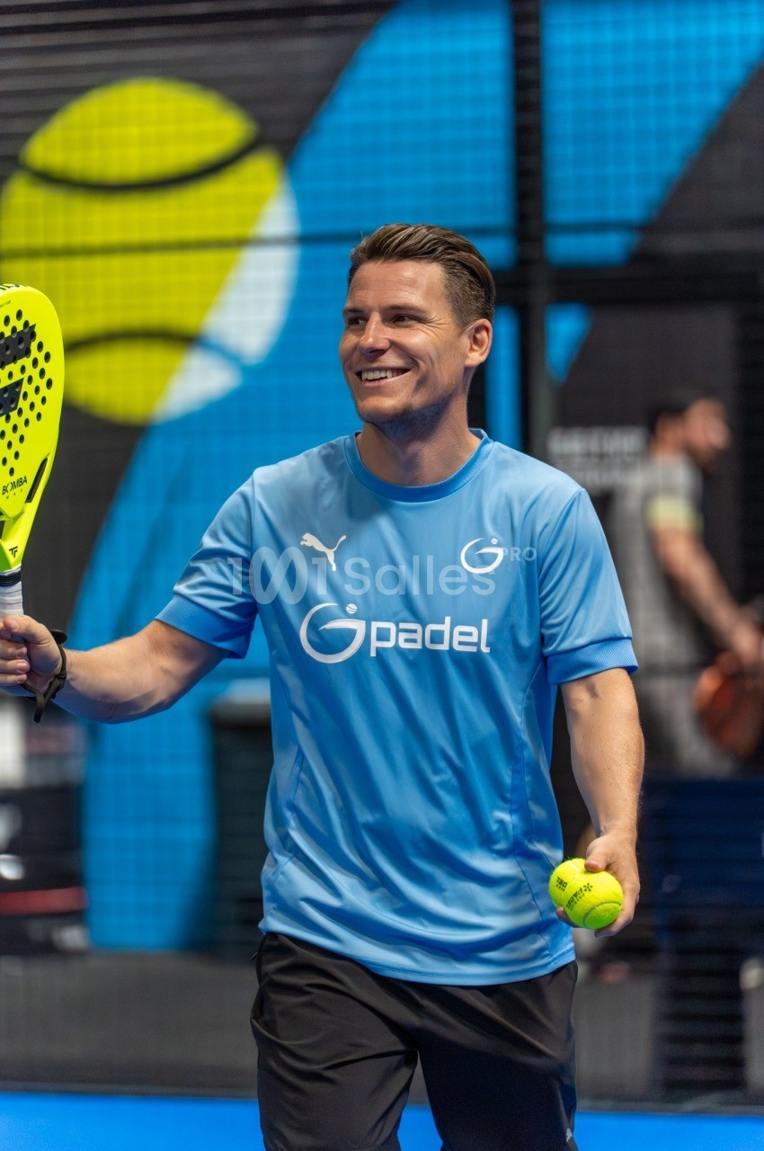 Homme tenant une raquette de padel et une balle jaune dans un club de sport couvert avec mur bleu.