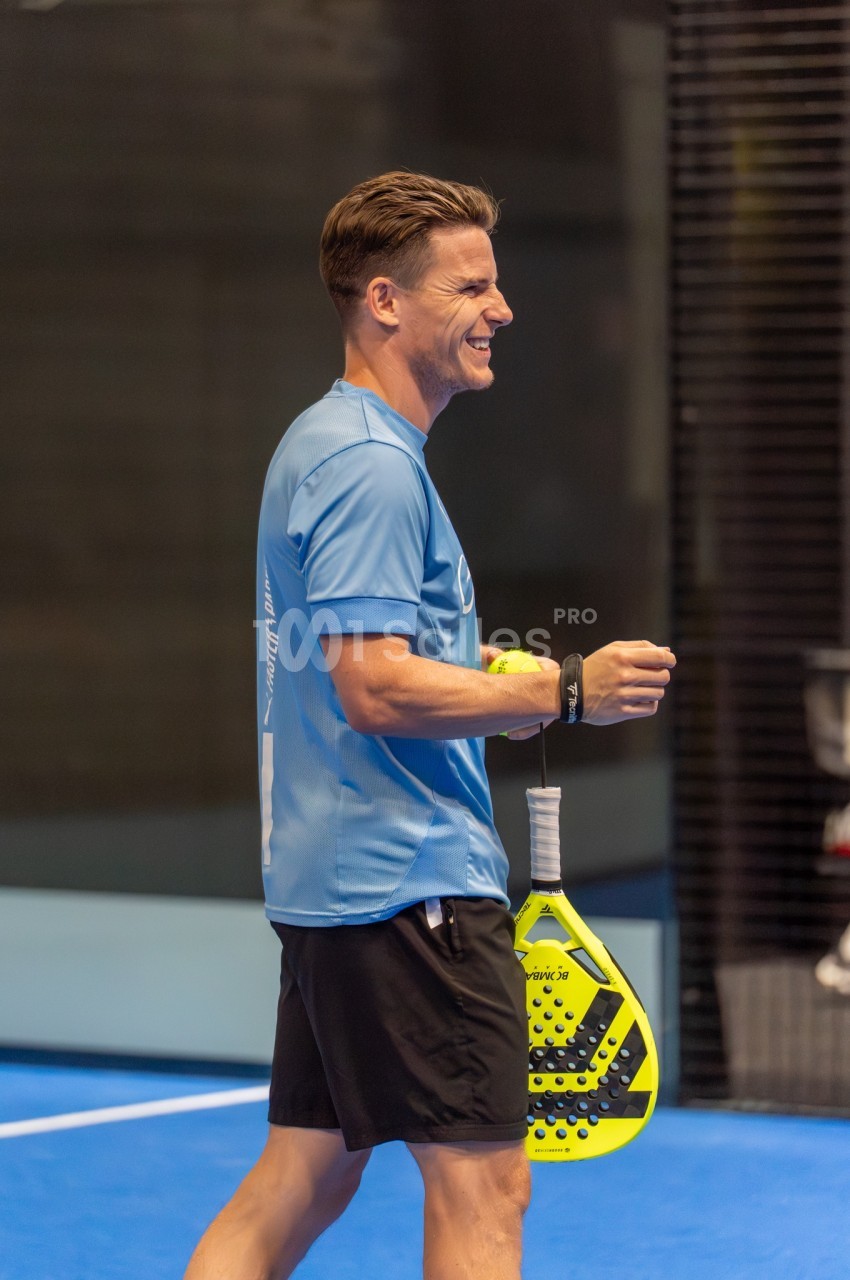 Moniteur en tenue de sport tenant une raquette jaune sur un terrain de squash aux murs bleu et noir.