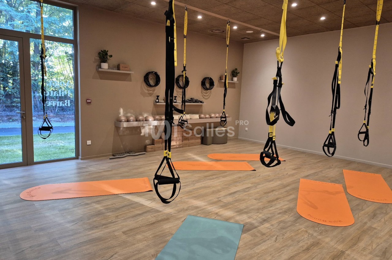 Salle de fitness avec équipements de suspension jaunes et noirs, tapis orange et bleu au sol, baies vitrées et mobilier de rangement.