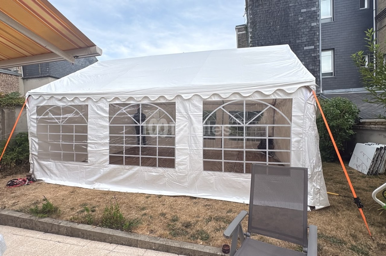 Chapiteau blanc rectangulaire avec parois transparentes installé en jardin, adossé à une bâtisse en pierre.